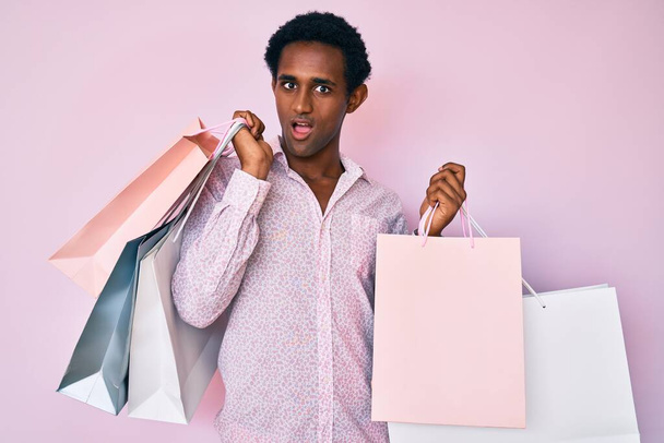 Africano guapo hombre sosteniendo bolsas de compras en la cara de choque, mirando escéptico y sarcástico, sorprendido con la boca abierta  - Foto, imagen