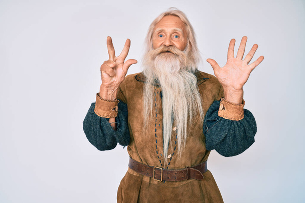 Vieil homme âgé aux cheveux gris et à la barbe longue portant un costume traditionnel viking montrant et pointant vers le haut avec les doigts numéro huit tout en souriant confiant et heureux.  - Photo, image