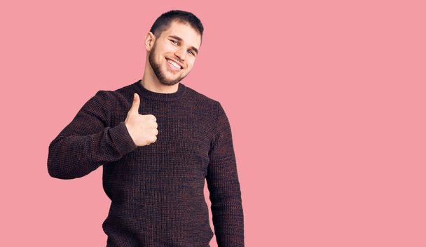 Young handsome man wearing casual sweater doing happy thumbs up gesture with hand. approving expression looking at the camera showing success.  - Photo, Image