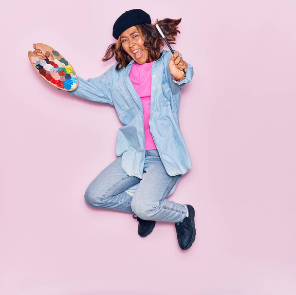 Jovem bela artista latino mulher vestindo boina francesa sorrindo feliz. Saltando com sorriso na pintura facial usando pincel e paleta sobre fundo rosa isolado - Foto, Imagem
