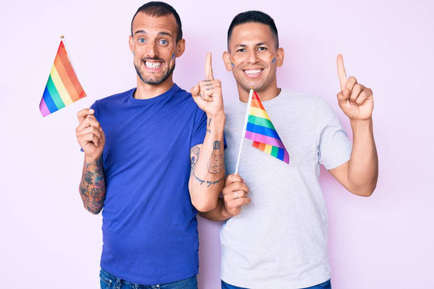 jong gay paar van twee mannen holding regenboog lgbtq vlaggen samen glimlachen met een idee of vraag wijzend vinger met gelukkig gezicht nummer een  - Foto, afbeelding