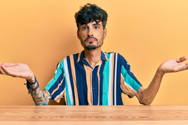 Young hispanic man wearing casual clothes sitting on the table clueless and confused expression with arms and hands raised. doubt concept.  - Photo, Image