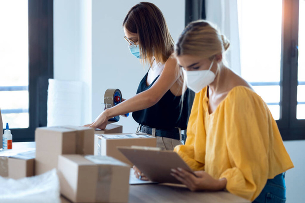 Shot of two beautiful freelance business women seller wearing a hygienic facial mask while checking product order while packing and sealing cardboard boxes in their startup small business.  - Foto, afbeelding