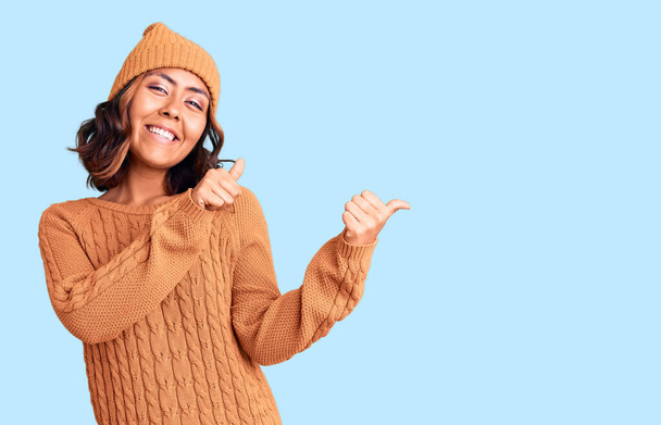 Nuori kaunis sekoitettu rotu nainen yllään villa pusero ja talvi hattu osoittaa takana käsi ja peukalot ylöspäin, hymyillen luottavainen  - Valokuva, kuva