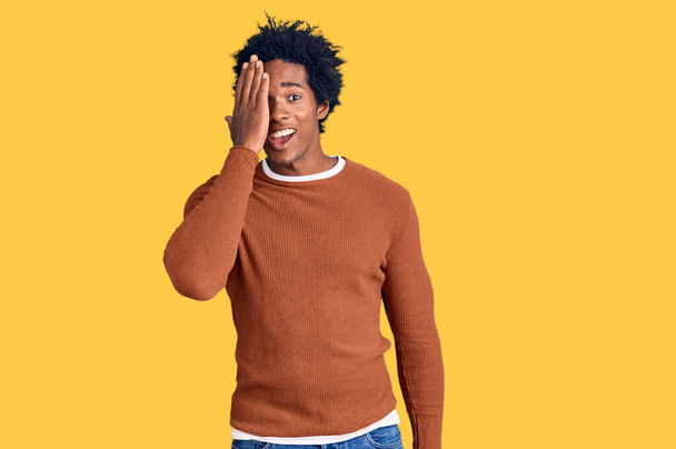Handsome african american man with afro hair wearing casual clothes covering one eye with hand, confident smile on face and surprise emotion.  - Photo, Image