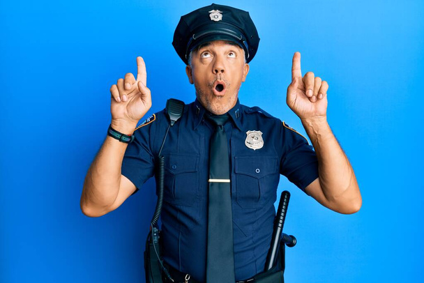 Knappe middelbare leeftijd volwassen man in politie-uniform verbaasd en verrast omhoog te kijken en te wijzen met vingers en opgeheven armen.  - Foto, afbeelding