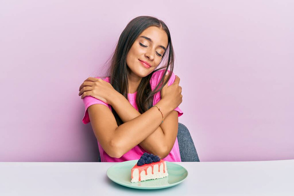 Молодая латиноамериканка ест чизкейк, сидя на столе, обнимая себя счастливой и позитивной, уверенно улыбаясь. любовь к себе и забота о себе  - Фото, изображение
