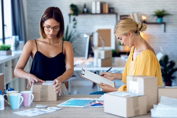 Shot of two beautiful freelance business women seller checking product order while packing and sealing cardboard boxes for delivery products ordered online to customers in their startup small business.  - Fotó, kép