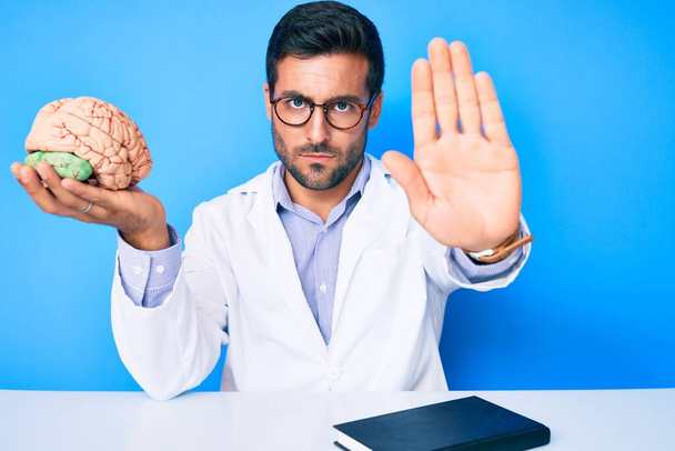 Young hispanic man sitting at the table wearing doctor coat holding brain with open hand doing stop sign with serious and confident expression, defense gesture  - Fotó, kép