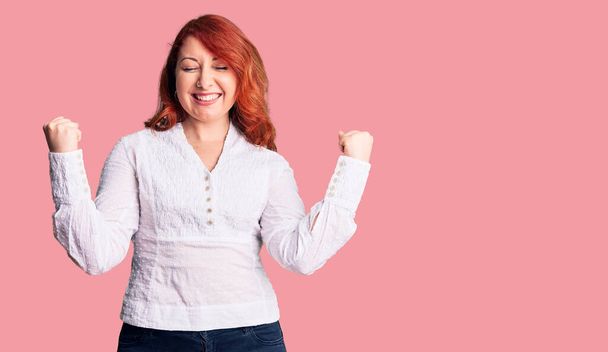 Young beautiful redhead woman wearing casual shirt very happy and excited doing winner gesture with arms raised, smiling and screaming for success. celebration concept.  - Foto, afbeelding