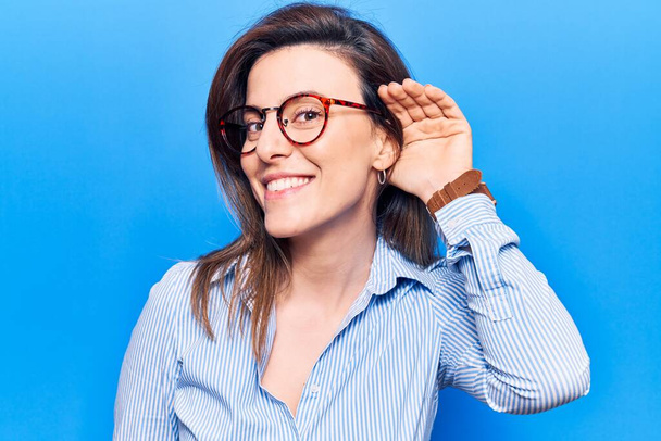 Young beautiful woman wearing business clothes and glasses smiling with hand over ear listening and hearing to rumor or gossip. deafness concept.  - Photo, Image