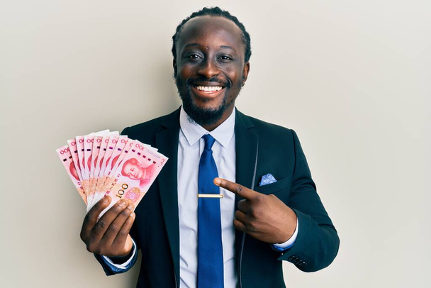 Handsome young black man wearing business suit holding 100 yuan banknotes smiling happy pointing with hand and finger  - 写真・画像
