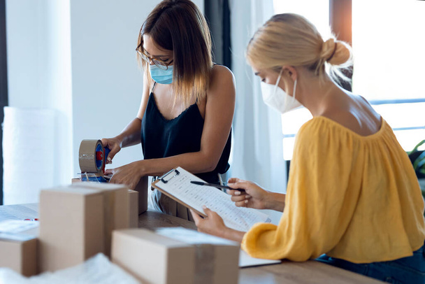 Shot of two beautiful freelance business women seller wearing a hygienic facial mask while checking product order while packing and sealing cardboard boxes in their startup small business.  - Fotografie, Obrázek