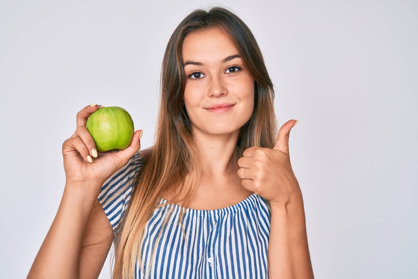 Beautiful caucasian woman holding green apple smiling happy and positive, thumb up doing excellent and approval sign  - Fotoğraf, Görsel