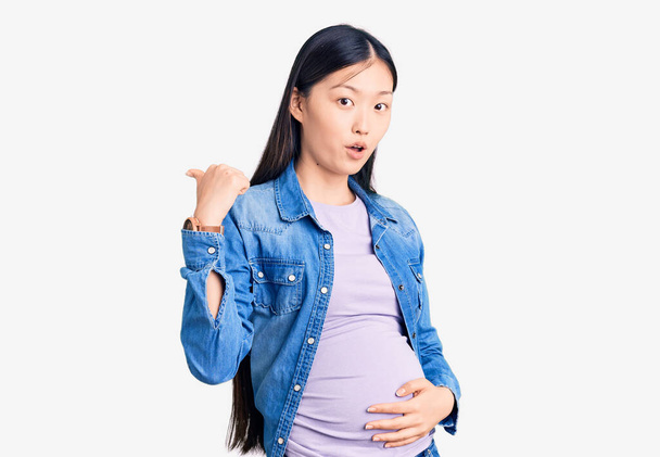 Νεαρή όμορφη Κινέζα έγκυος περιμένει μωρό έκπληκτος δείχνοντας με το χέρι δάχτυλο στο πλάι, ανοιχτό στόμα κατάπληκτη έκφραση.  - Φωτογραφία, εικόνα