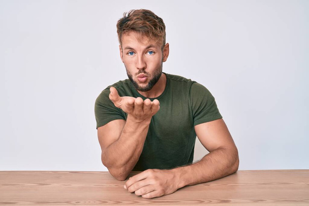 Young caucasian man wearing casual clothes sitting on the table looking at the camera blowing a kiss with hand on air being lovely and sexy. love expression.  - Photo, Image