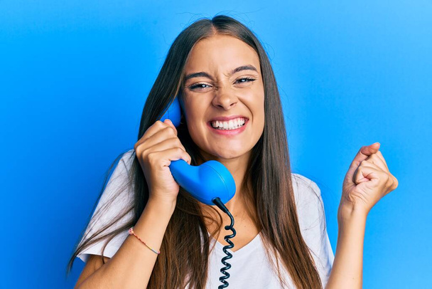 Young hispanic woman holding vintage telephone screaming proud, celebrating victory and success very excited with raised arm  - Фото, изображение