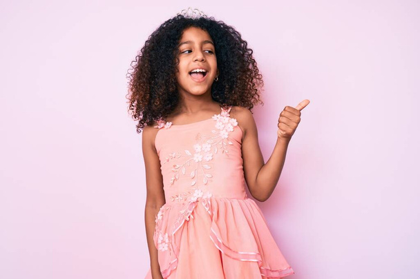Africké americké dítě s kudrnatými vlasy nosí princeznu korunu ukazující palec nahoru na stranu s úsměvem šťastný s otevřenými ústy  - Fotografie, Obrázek