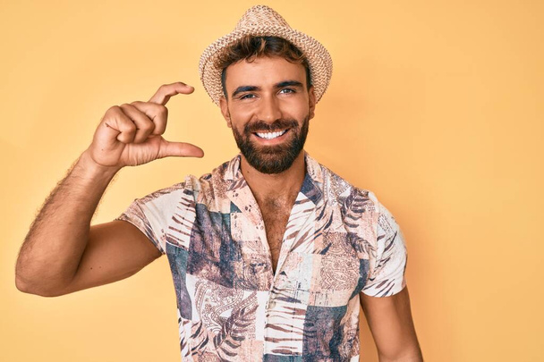 Jonge Spaanse man met zomerhoed lachend en zelfverzekerd gebaar met de hand die klein bord met vingers kijkt en de camera doet. meetconcept.  - Foto, afbeelding