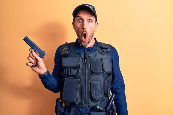 Handsome policeman wearing uniform and bulletprof holding gun over yellow background scared and amazed with open mouth for surprise, disbelief face - Photo, Image