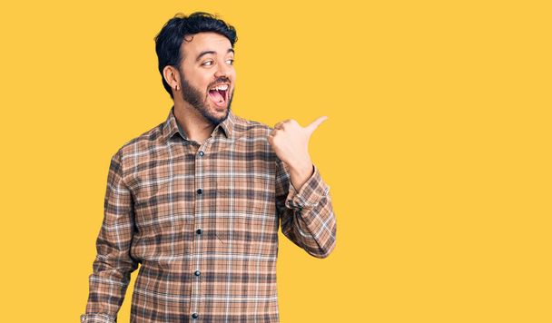 Jonge Spaanse man draagt casual kleding glimlachend met een vrolijk gezicht en wijzend naar de zijkant met duim omhoog.  - Foto, afbeelding
