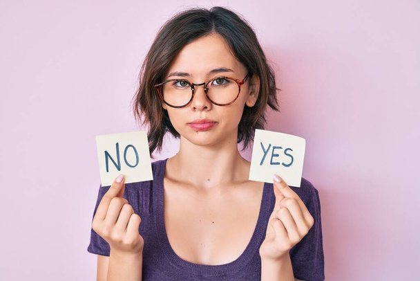 Young beautiful girl holding yes and no reminder relaxed with serious expression on face. simple and natural looking at the camera.  - Foto, imagen