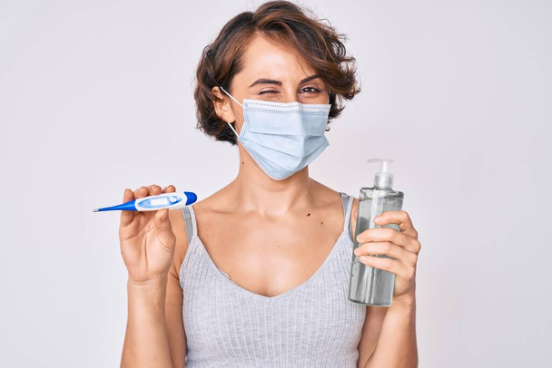 Fiatal spanyol nő orvosi maszkot visel, kézfertőtlenítő gél és hőmérő kacsintás néz a kamera szexi kifejezés, vidám és boldog arc.  - Fotó, kép