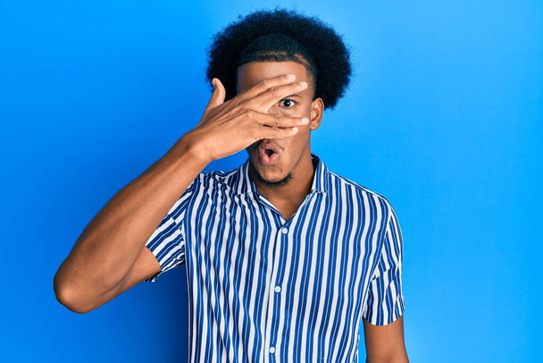Afrikkalainen amerikkalainen mies afro hiukset yllään rento vaatteita kurkistaa shokki peittää kasvot ja silmät käsin, katselee sormien läpi peloissaan  - Valokuva, kuva
