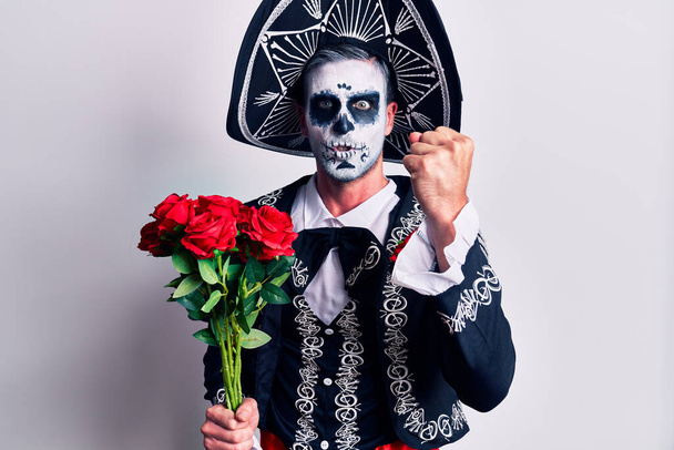 Νεαρός άνδρας φορώντας μεξικάνικη μέρα της νεκρής στολής κρατώντας λουλούδια ενοχλημένος και απογοητευμένος φωνάζοντας από θυμό, φωνάζοντας τρελά από θυμό και σηκώνοντας το χέρι  - Φωτογραφία, εικόνα