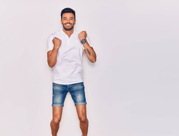 Young handsome hispanic man wearing casual clothes smiling happy. Jumping with smile on face celebrating with fists up over isolated white background - Valokuva, kuva