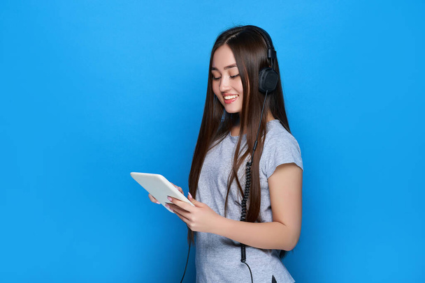 Smiling young beautiful asian girl listening to music on headphones and holds tablet on blue background. - 写真・画像