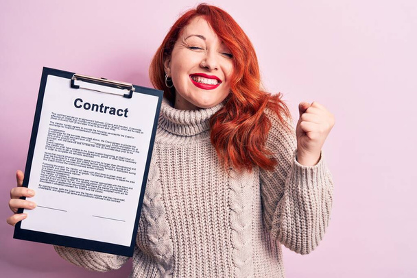 Young redhead woman holding clipboard with contract document paper over pink background screaming proud, celebrating victory and success very excited with raised arm - Foto, imagen