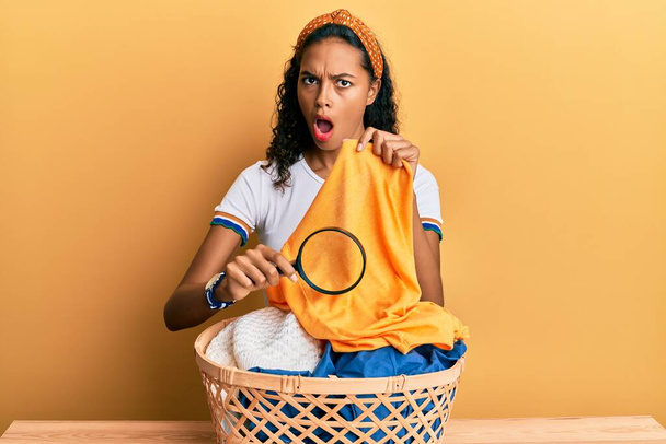 jong Afrikaans amerikaans meisje houden vergrootglas op zoek naar vlek op kleding in shock gezicht, op zoek sceptisch en sarcastisch, verrast met open mond  - Foto, afbeelding