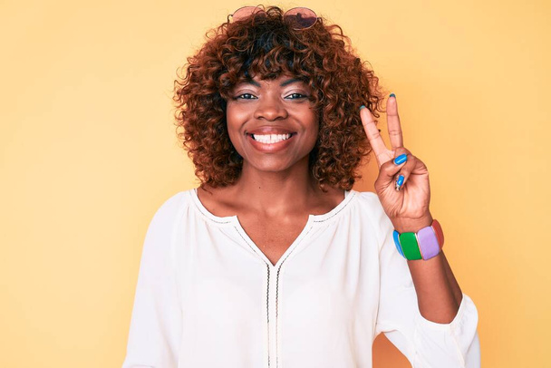 Jeune femme afro-américaine portant des vêtements décontractés souriant avec un sourire heureux clin d'oeil à la caméra faisant signe de victoire. numéro deux.  - Photo, image