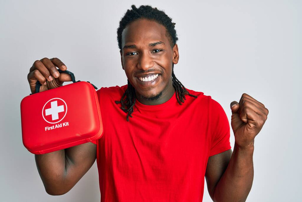African american man with braids holding first aid kit screaming proud, celebrating victory and success very excited with raised arm  - Foto, Bild