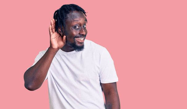 Ein junger afrikanisch-amerikanischer Mann mit Zöpfen in einem lässigen weißen T-Shirt, der lächelnd mit der Hand über dem Ohr Gerüchten oder Klatsch lauscht. Konzept für Gehörlosigkeit.  - Foto, Bild