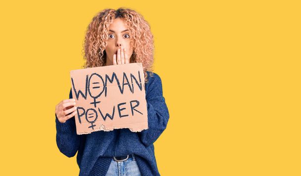 Young blonde woman with curly hair holding we need a change banner covering mouth with hand, shocked and afraid for mistake. surprised expression  - 写真・画像