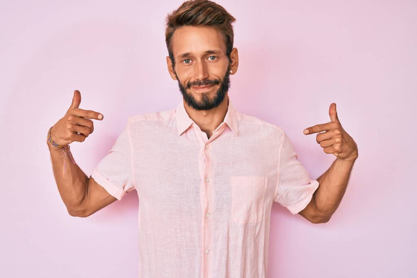 Όμορφος καυκάσιος άντρας με γενειάδα φοράει ροζ πουκάμισο που δείχνει σίγουρος με χαμόγελο στο πρόσωπο, δείχνοντας τον εαυτό του με τα δάχτυλα του περήφανα και χαρούμενα.  - Φωτογραφία, εικόνα