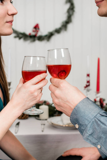 A close picture of young spouses clinking glasses and looking at each other. Beautiful couple sitting at the laid table for thanksgiving day, holding glass of wine in the hands. - Photo, Image