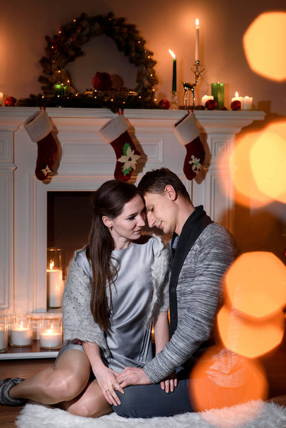 A lovely couple cuddling gently on a white furry carpet near a christmas decorated fireplace, sunny bunnies playing on a picture. Young couple sitting on floor at burning fireplace. - Foto, imagen