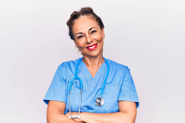 Middle age brunette nurse woman wearing uniform and stethoscope over white background happy face smiling with crossed arms looking at the camera. Positive person. - Zdjęcie, obraz