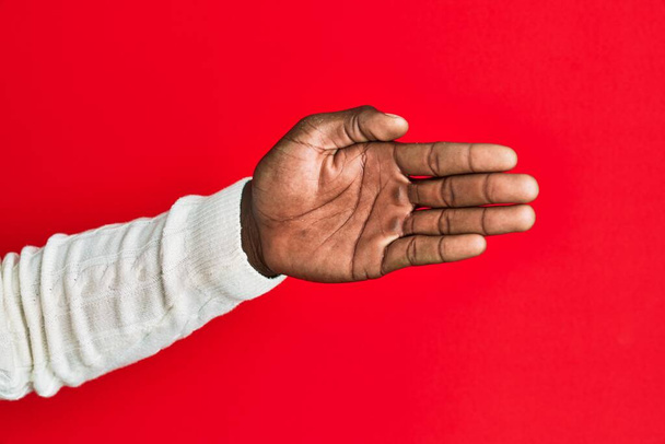 Brazo y mano del joven afroamericano negro sobre fondo rojo aislado estirándose y alcanzando con la mano abierta para el apretón de manos, mostrando la palma  - Foto, imagen