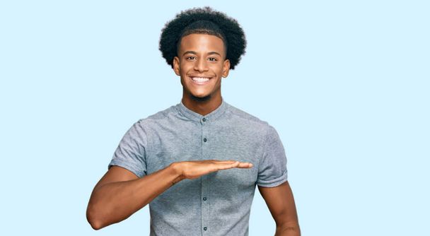 Afro-Amerikaanse man met afrohaar in casual kleding gebaren met handen tonen grote en grote maat teken, maatregel symbool. glimlachend kijkend naar de camera. meetconcept.  - Foto, afbeelding