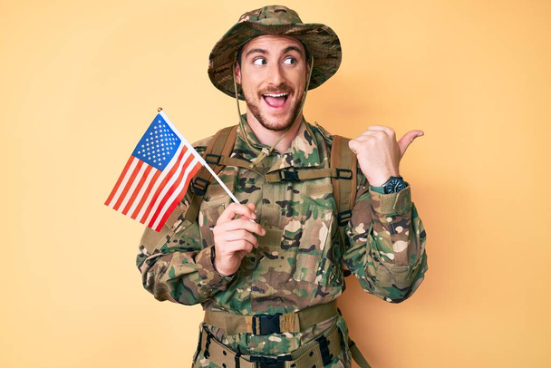 Joven hombre caucásico con camuflaje uniforme del ejército sosteniendo la bandera de EE.UU. señalando el pulgar hacia el lado sonriendo feliz con la boca abierta  - Foto, imagen