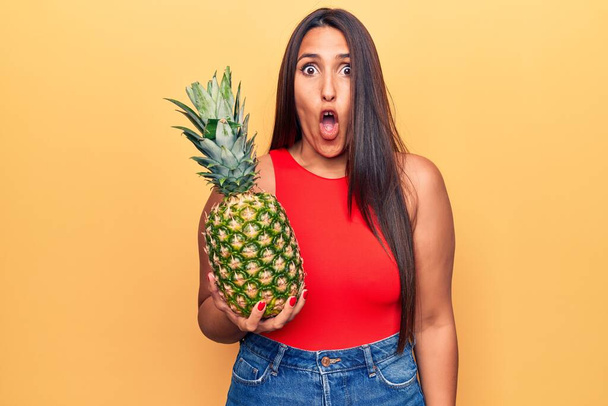 Nuori kaunis brunette nainen tilalla ananas peloissaan ja hämmästynyt suu auki yllätys, epäusko kasvot  - Valokuva, kuva