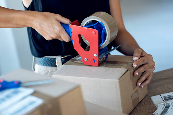 Close-up of young woman hands holding packing machine and sealing cardboard boxes with duct tape to delivery products ordered online to customers in her startup small business.  - Foto, Bild