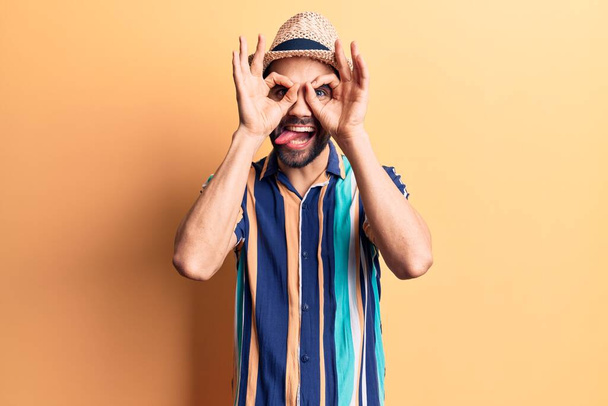 Young handsome man with beard wearing summer hat and shirt doing ok gesture like binoculars sticking tongue out, eyes looking through fingers. crazy expression.  - Fotoğraf, Görsel