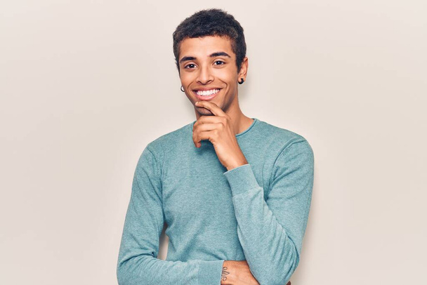 カジュアルな服を着た若いアフリカ系アメリカ人男性が、交差する腕と顎の手でカメラに自信を持って笑っている。ポジティブな.  - 写真・画像