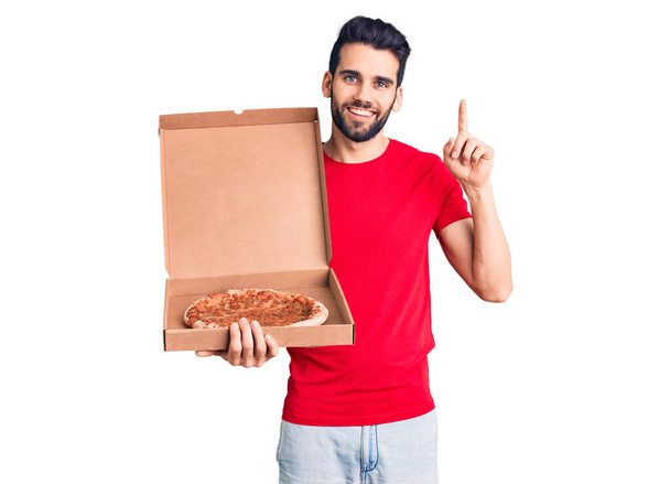 Joven hombre guapo con barba sosteniendo cartón de entrega con pizza italiana sorprendido con una idea o pregunta señalando el dedo con la cara feliz, número uno  - Foto, imagen