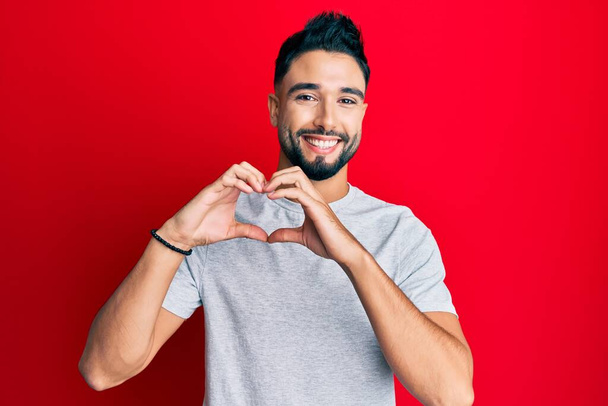 Jeune homme à la barbe portant un t-shirt blanc décontracté souriant en amour faisant forme de symbole de coeur avec les mains. concept romantique.  - Photo, image